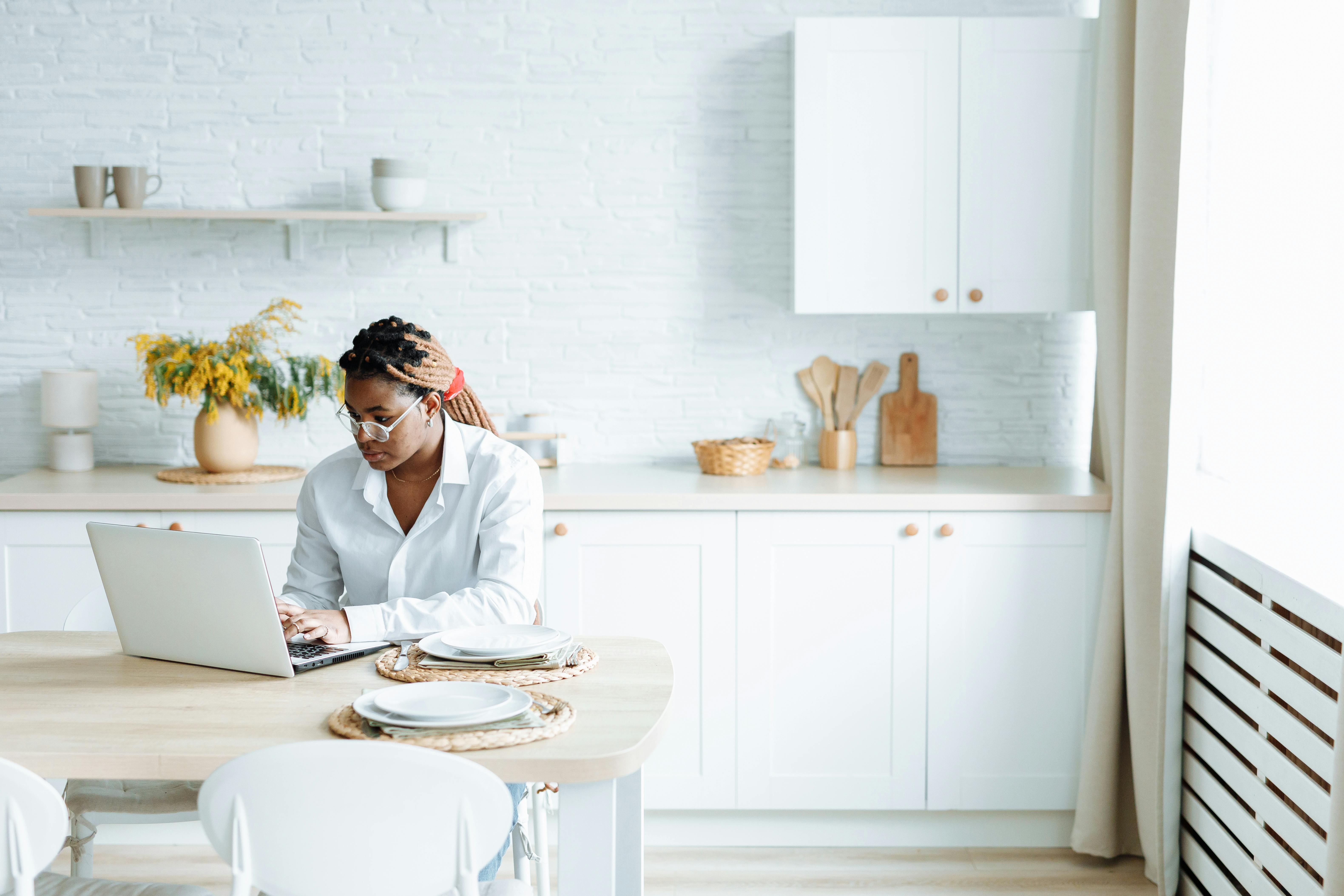 A black woman types on her laptop in a bright, modern kitchen, embodying work-from-home lifestyle.